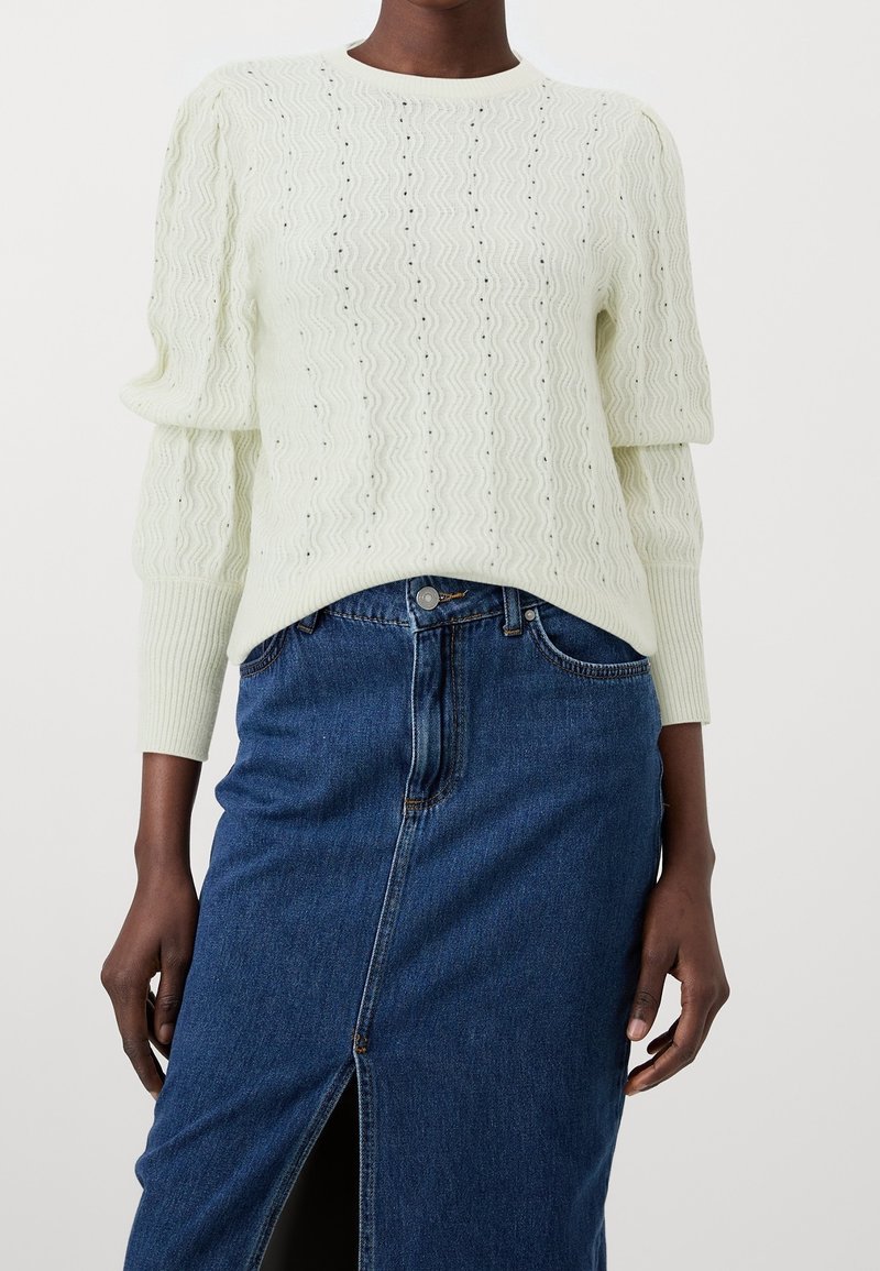 Femme portant un pull blanc à manches longues texturé avec des épaules bouffantes, associé à une jupe en jean bleue taille haute avec une fente à l'avant.