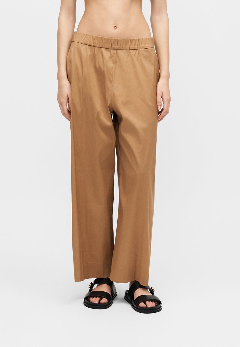 Λεπτή μέση που φοράει φαρδιά παντελόνια με ψηλή μέση σε καμηλό χρώμα με ελαστική μέση και μαύρα σανδάλια ανοιχτών δαχτύλων σε λευκό φόντο.