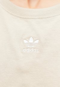 Bézs pulóver bordázott nyakkivágással, bal oldalon fehér hímzett Adidas háromleveleű lóhere logóval. Puha pamut textúra.