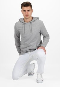 Haut en coton gris avec une poche frontale, un capuchon à cordon et des manches longues, associé à un pantalon blanc avec des coutures jaunes et des baskets blanches.