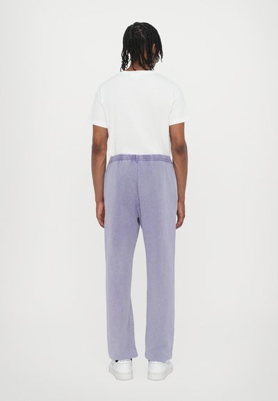Purple sweatpants with a relaxed fit, featuring an elastic waistband, worn with a white t-shirt and white sneakers. Smooth textile finish.