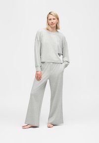 Γυναίκα που στέκεται ξυπόλητη φορώντας άνετο ανοιχτογκρι μακρυμάνικο μπλουζάκι και παντελόνι με φαρδιά πόδια μπροστά σε απλό λευκό φόντο.