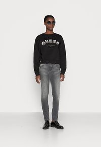 Svart kort sweatshirt med metallisk "Guess California" bokstavering, tillsammans med grå slim-fit jeans och glansiga svarta skor.