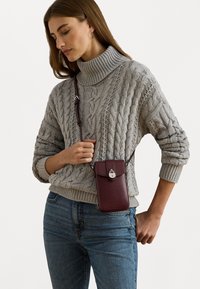 Tailliertes graues Kabelstrickpullover, hoher Kragen, kombiniert mit einer bordeauxfarbenen Umhängetasche für das Handy, die einen Druckverschluss und einen verstellbaren Tragegurt hat.