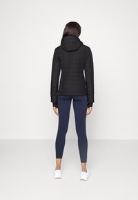 Svart pufferjacka med huva, med kviltad textur. Tillsammans med marinblå tights och vita träningsskor.