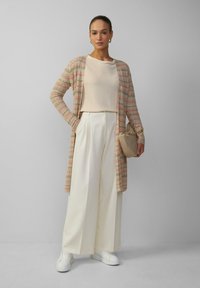 Langer, offener Cardigan mit einem wellenförmigen Muster in Beige, Rosa und Orange; kombiniert mit einem hellen Top, weiten weißen Hosen und weißen Sneakers.