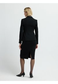 Blazer negro ajustado con un diseño ceñido, combinado con una falda lápiz a la altura de la rodilla que presenta una abertura en la parte posterior, acompañado de medias negras transparentes y tacones.