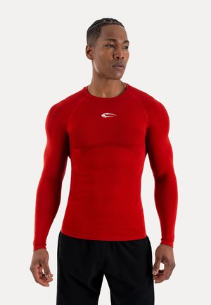 Muskularny mężczyzna z zaplecionymi włosami, ubrany w dopasowaną, czerwoną, długorękawową sportową koszulkę i czarne spodnie, zwrócony lekko w prawo.