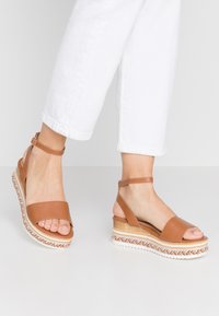 Sandales compensées en cuir marron avec une large bride au niveau des orteils et une bride ajustable à la cheville. Texture tressée sur une semelle plate, combinées avec un jean blanc.