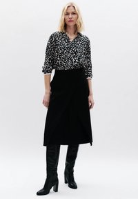 Zwarte wikkelrok in combinatie met een zwart-wit gespikkelde blouse. Model draagt kniehoog zwarte haklaarzen. De textuur van de stof contrasteert met de ontwerpen.