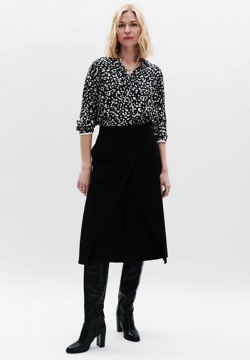 Zwarte wikkelrok in combinatie met een zwart-wit gespikkelde blouse. Model draagt kniehoog zwarte haklaarzen. De textuur van de stof contrasteert met de ontwerpen.