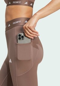Hnědé sportovní legíny s boční kapsou na chytrý telefon. Vybaveny vysokým pasem s logem "TECHFIT" a elastickým materiálem.