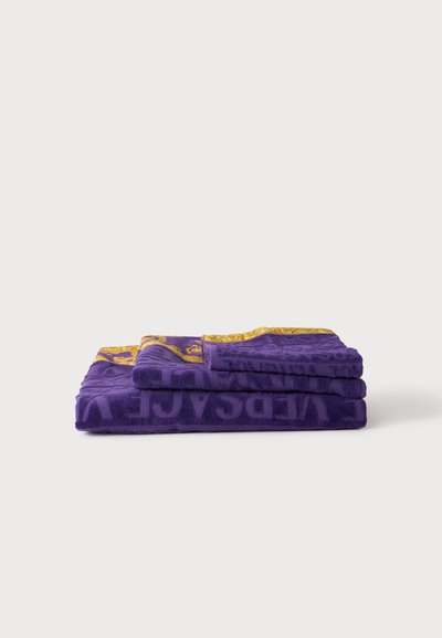 Pile de trois serviettes violettes pliées avec des lettres en relief et des bordures ornées de motifs jaunes et dorés sur un fond blanc uni.