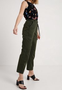 Top senza maniche nero con stampa di farfalle colorate, abbinato a pantaloni cargo verde oliva con tasche e una vestibilità sartoriale. Sandali neri con tacco a blocco.