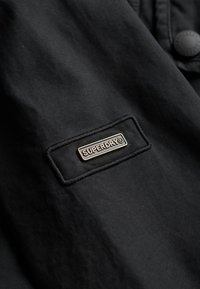 Sort jakke i stof med en rektangulær applikation med "SUPERDRY" logoet i gyldne bogstaver og omgivende syning. Knapdetalje synlig.