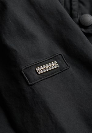Black jacket fabric featuring a rectangular patch with the "SUPERDRY" logo in gold lettering and outlined stitching. Button detail visible.