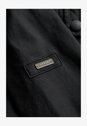 Tissu de veste noire avec une patch rectangulaire arborant le logo "SUPERDRY" en lettres dorées et avec une couture en surpiqûre. Détail de boutons visible.