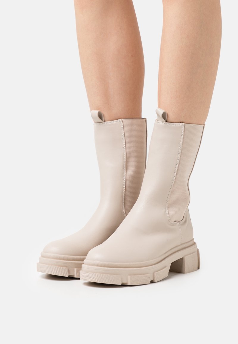Bottes basses en cuir beige avec une semelle épaisse, des panneaux latéraux élastiques et des languettes pour un enfilage facile. Texture lisse, design minimaliste.