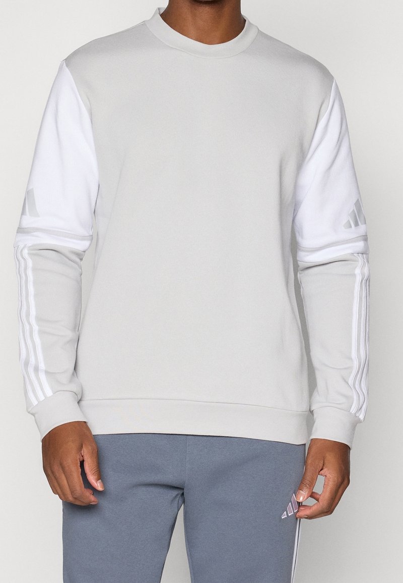 Homme portant un sweat-shirt gris clair avec des manches blanches et des bandes sur les bras, associé à un pantalon bleu-gris avec un petit logo près de la poche.