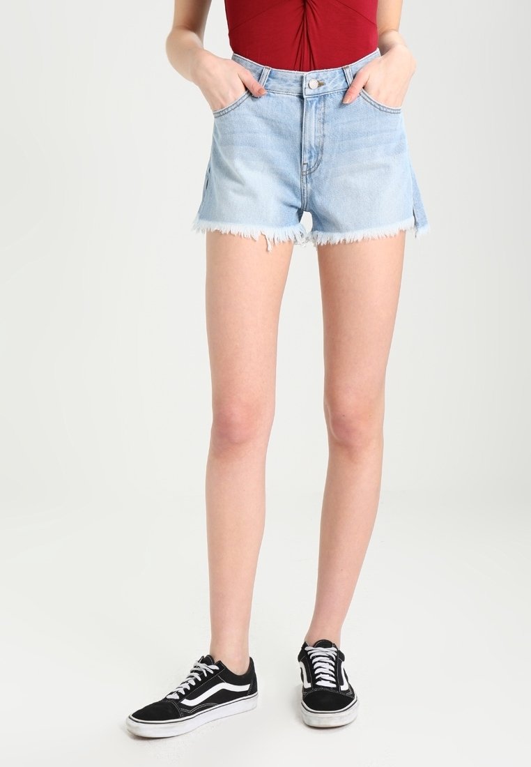 Femme portant un short en denim bleu clair effiloché, un haut rouge et des baskets noires et blanches, debout les mains dans les poches sur un fond blanc.