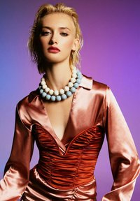 Blonde woman with wet hair wearing a pink satin dress and large blue and white beaded necklace against a purple gradient background.