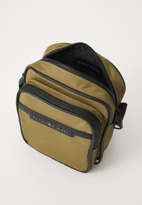 Borsa in nylon verde oliva con più scomparti con zip, rifiniture nere e un'etichetta con il logo. Zip robusta con tirante e dettagli di cucitura a contrasto.