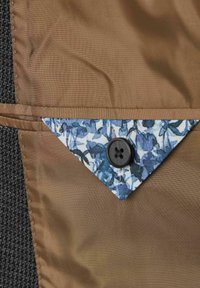 Brauner Stoff mit einem floralen Taschen Detail in Blau und Weiß, versehen mit einem schwarzen Knopf. Glatte Textur mit einer maßgeschneiderten Verarbeitung.