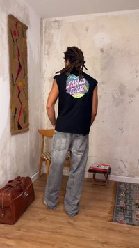 Άτομο με dreadlocks που φοράει μαύρο αμάνικο μπλουζάκι "Santa Cruz" και γκρι τζιν στέκεται σε ένα φθαρμένο δωμάτιο με ξύλινο πάτωμα και σκορπισμένα έπιπλα.
