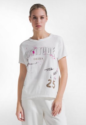 Giovane donna con capelli chiari raccolti, che indossa una t-shirt bianca con grafica, testo e numeri, in piedi davanti a uno sfondo bianco uniforme.