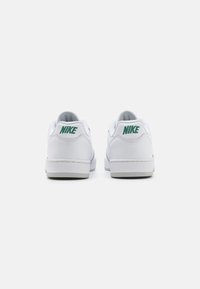 Fehér sportcipő sima bőr felsőrésszel, párnázott nyakkal és zöld Nike logóval a sarkán. A talpa szürke, minimális barázdákkal.