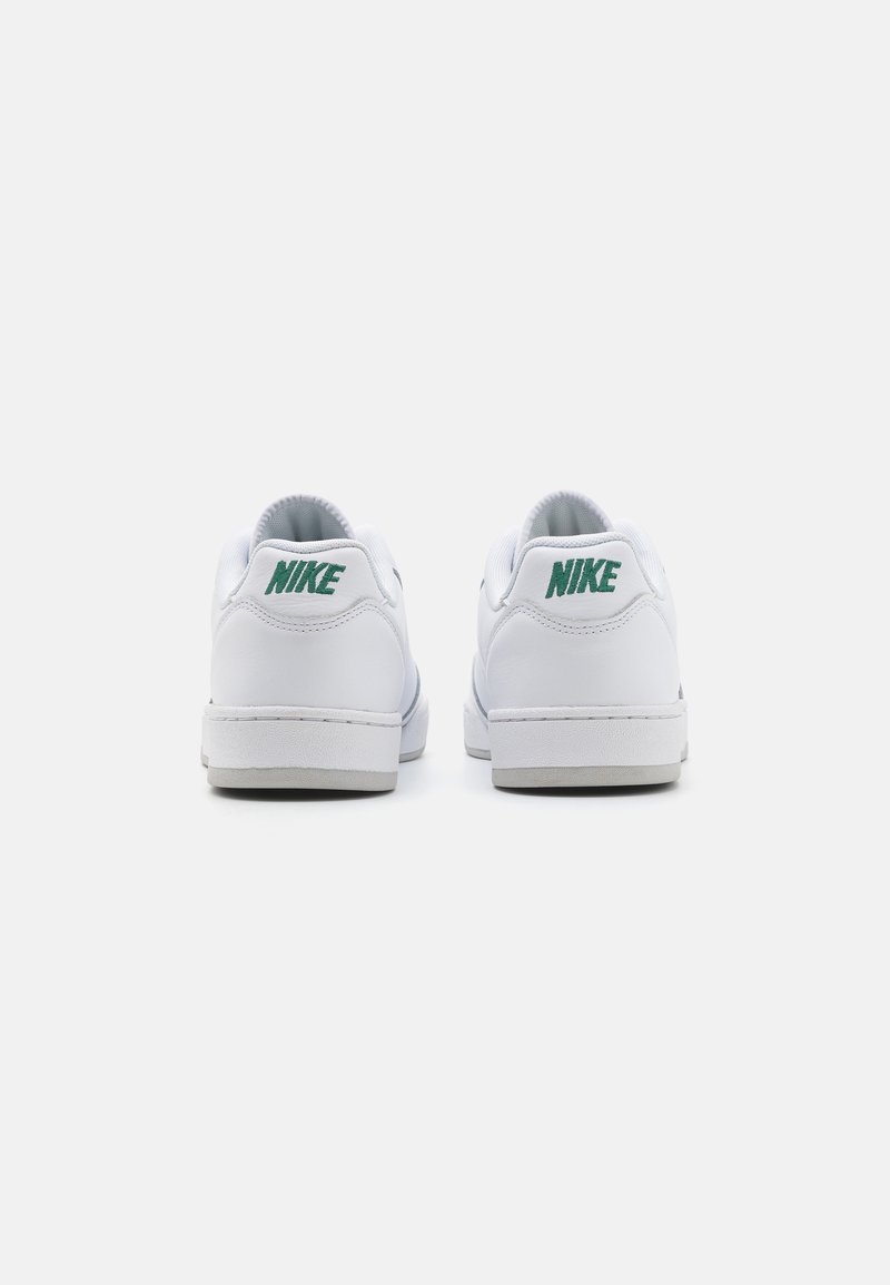 Baskets blanches avec un dessus en cuir lisse, un col rembourré et un logo Nike vert au niveau du talon. La semelle extérieure est grise avec des motifs de semelle minimalistes.