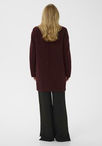 Burgundska dolga jopica s ravnim krojem, teksturiranim pletenjem in spušenimi rameni, v kombinaciji s širokimi črnimi hlačami.