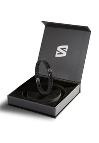 Black metal chain bracelet displayed in a black box with a soft, round cushion. Box features a white logo on the inner lid.