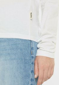 Langarmweißes Baumwolloberteil mit einem Etikett "SENSES" an der Seite, kombiniert mit hellblauen Jeans. Glatte Textur und einfaches Design.