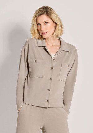 Blonde Frau mit kurzen Haaren, die eine beige Jacke mit Knöpfen und zwei Brusttaschen und passende Hose trägt, steht mit den Händen in den Taschen.