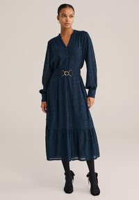 Navyblaues, gestuftes Kleid mit V-Ausschnitt, langen Ärmeln, strukturiertem Stoff und goldener Taillenschließe. Kombiniert mit schwarzen Strumpfhosen und Absatzschuhen.