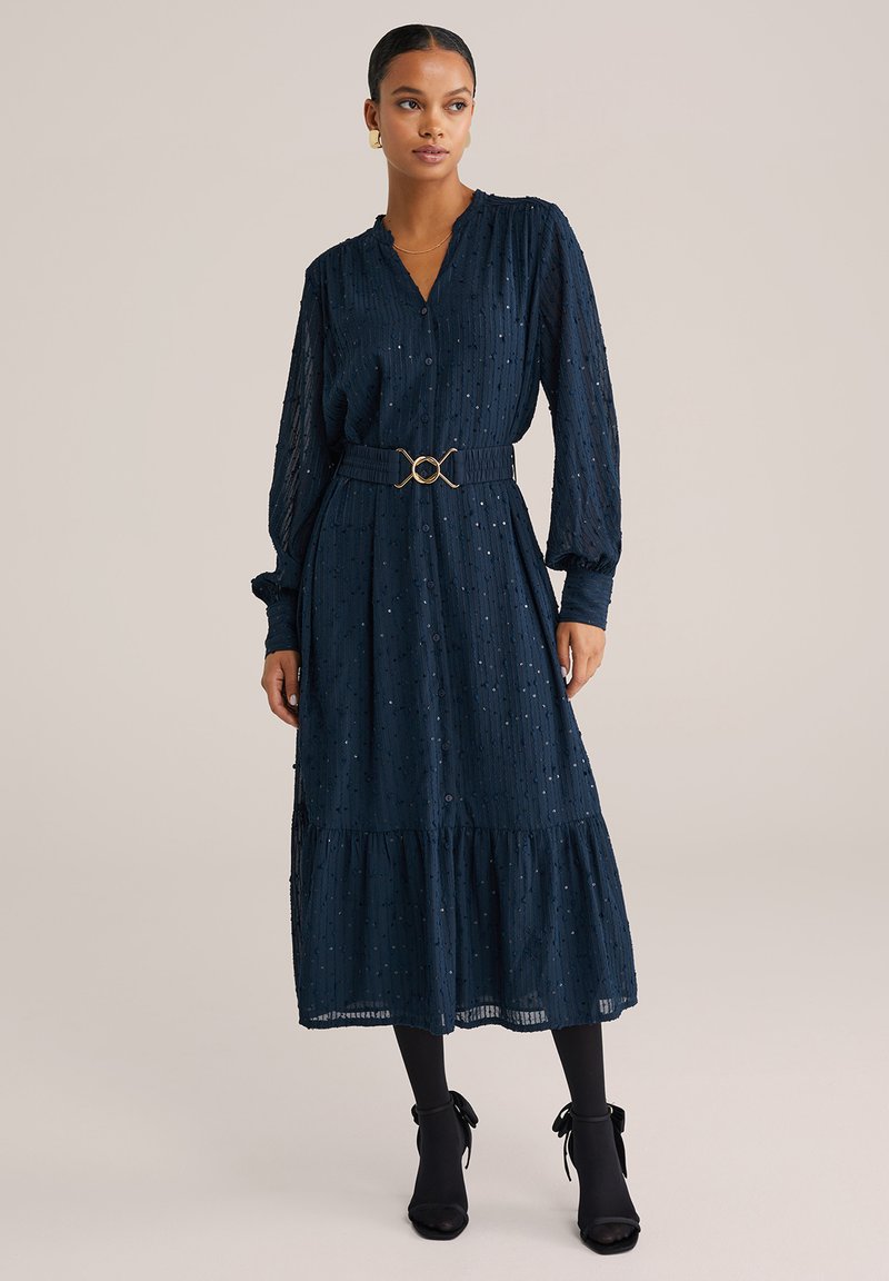 Navyblaues, gestuftes Kleid mit V-Ausschnitt, langen Ärmeln, strukturiertem Stoff und goldener Taillenschließe. Kombiniert mit schwarzen Strumpfhosen und Absatzschuhen.