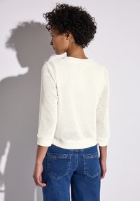 Pull à mailles blanches avec des manches trois-quarts et un motif texturé, associé à un jean bleu avec deux poches arrière.