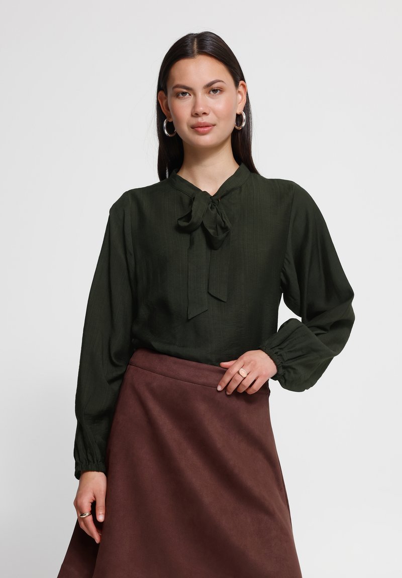 Femme portant un chemisier vert foncé à manches longues avec un nœud au col et une jupe marron taille haute, debout devant un fond uni.