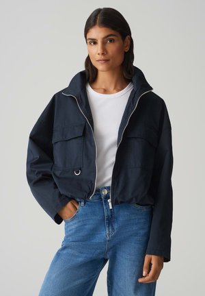 Gekürzte marineblaue Jacke mit zwei Fronttaschen, einem Reißverschluss und einem Stehkragen, getragen über einem weißen Oberteil und blauen Jeans.