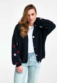 Žena s dlhými vlnitými vlasmi má na sebe čierny kardigán s červenými a ružovými hviezdami, biele tričko a svetlomodré rifle na jednobielom pozadí.