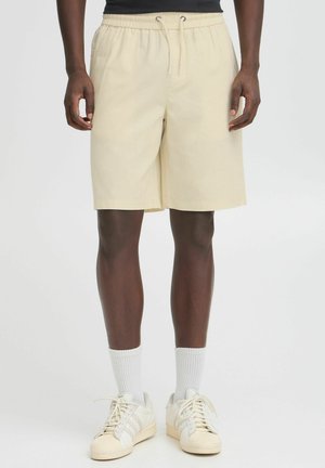 Shorts beige avec une taille élastique et un cordon de serrage, dotés d'une coupe ample et d'une texture lisse, associés à des baskets blanches et des chaussettes.