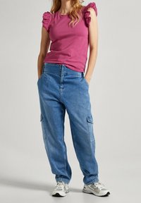 Różowa bawełniana koszulka z falbaniastymi koronkowymi rękawami, zestawiona z jasnoniebieskimi spodniami cargo z frontowymi kieszeniami i luźnym krojem. Białe sportowe sneakersy.