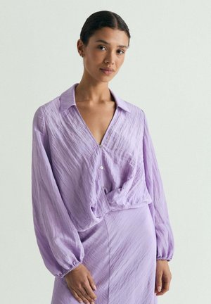 Mujer que lleva una blusa lavanda texturizada con mangas largas y una falda de talle alto a juego sobre un fondo claro liso.