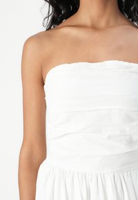 Bílé šaty bez ramínek s texturovaným živůtkem se shromážděnými detaily a splývavou sukní, vyrobené z lehké látky.