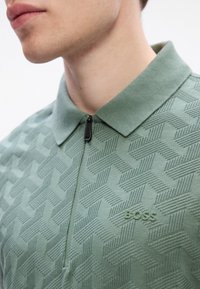 Närbild av en person som har på sig en grön mönstrad zip-up polo-tröja med en upphöjd "BOSS"-logotyp på bröstet.
