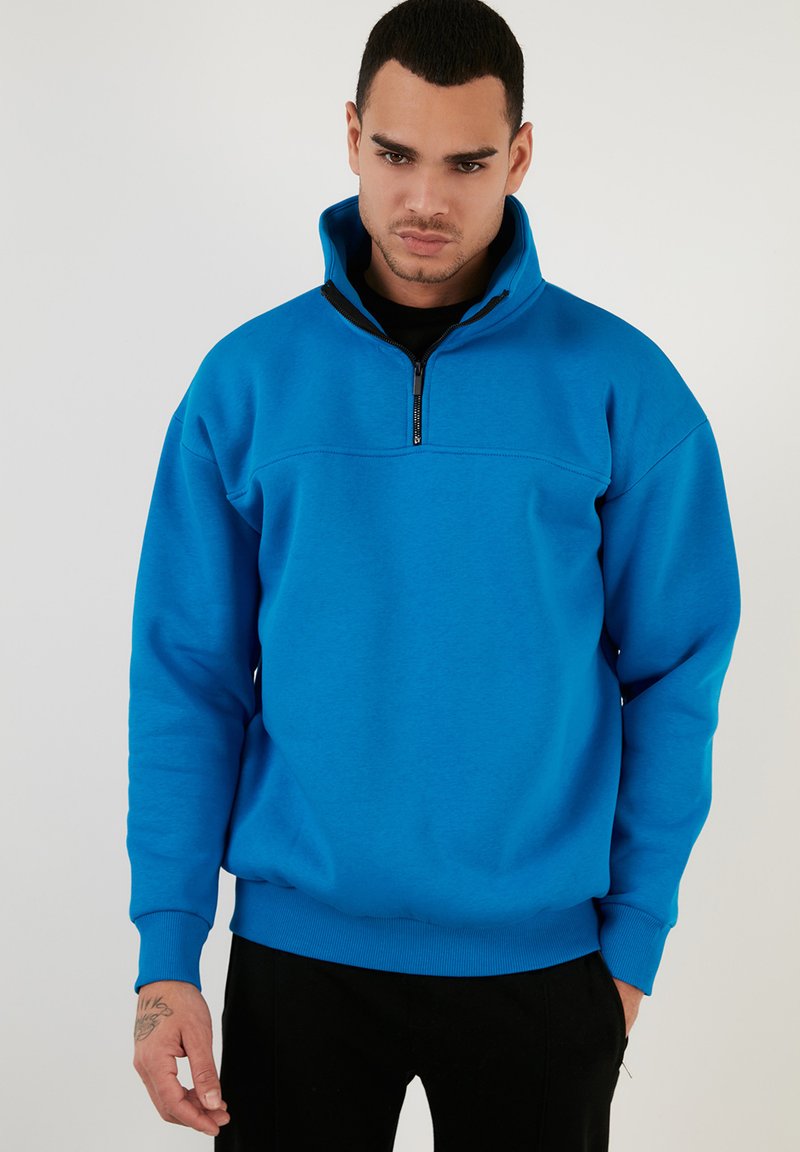 Sudadera de forro polar azul con cuello de media cremallera, mangas largas y puños acanalados. Presenta una textura suave y un diseño de corte relajado.