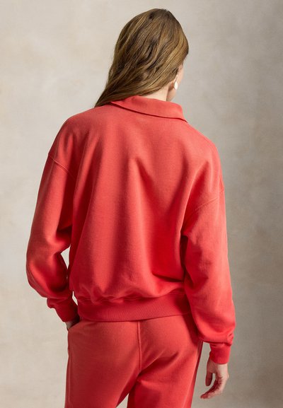 Mujer de cabello largo y castaño de espaldas, vestida con sudadera rosa coral y pantalones a juego, contra un fondo neutro.