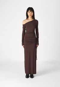 Langes, figurbetontes braunes Kleid mit einem schulterfreien Ärmel, strukturiertem Stoff und verstellbarem seitlichem Rüschendetail, getragen mit schwarzen Knöchelstiefeln.