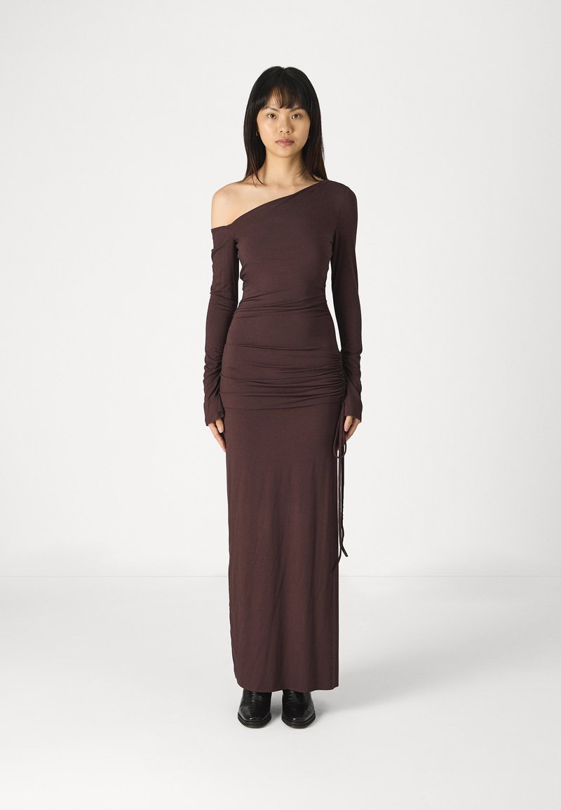 Langes, figurbetontes braunes Kleid mit einem schulterfreien Ärmel, strukturiertem Stoff und verstellbarem seitlichem Rüschendetail, getragen mit schwarzen Knöchelstiefeln.